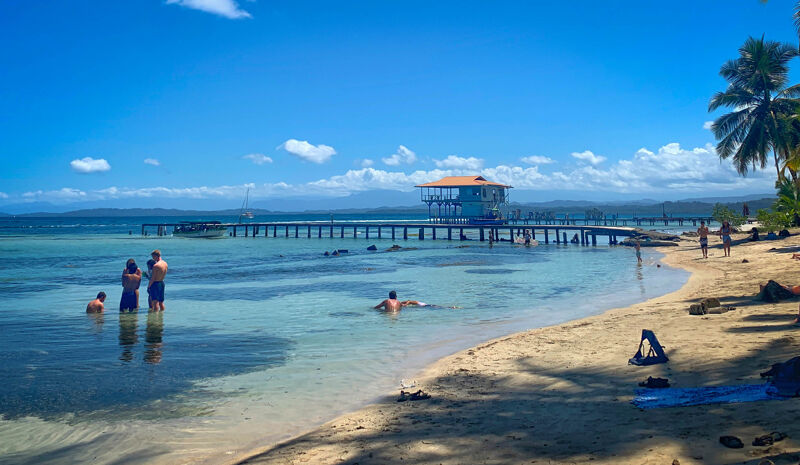 The image shows a beautiful beach scene on a sunny day. The water is clear and turquoise, and there are several people enjoying the water. There is a pier that extends into the water, and a building at the end of the pier. The beach is sandy, and there are palm trees in the background.
